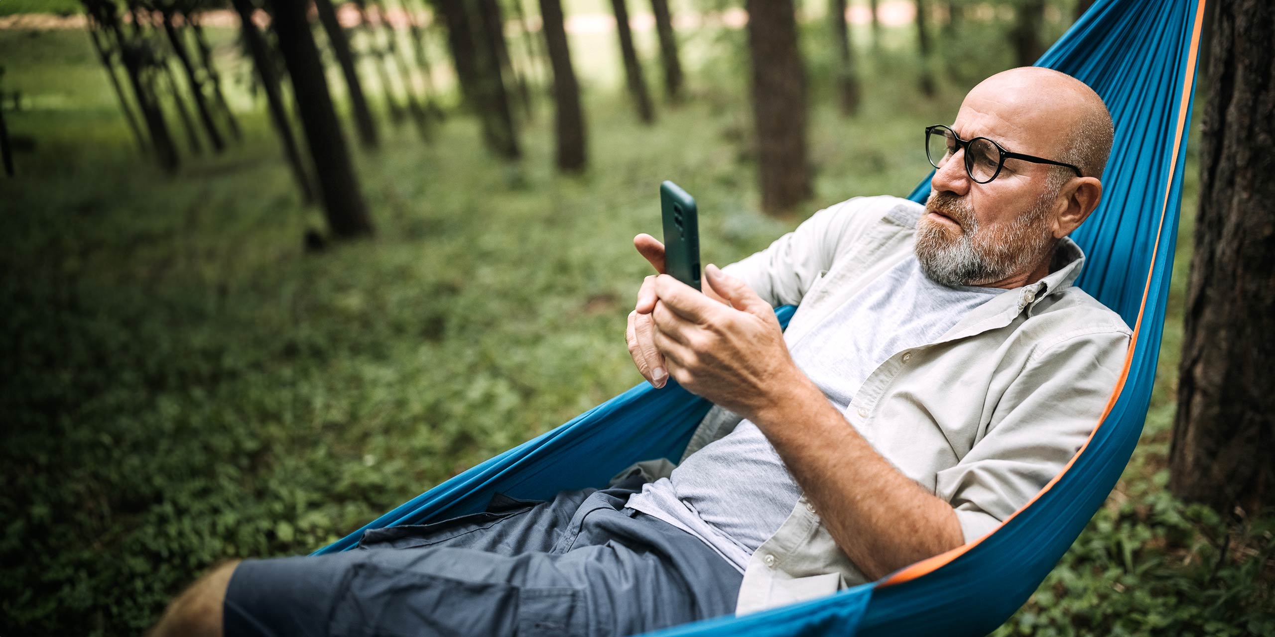 Ein entspannter Mann mit Bart liegt in einer blauen Hängematte im Wald und ist in sein Smartphone vertieft.