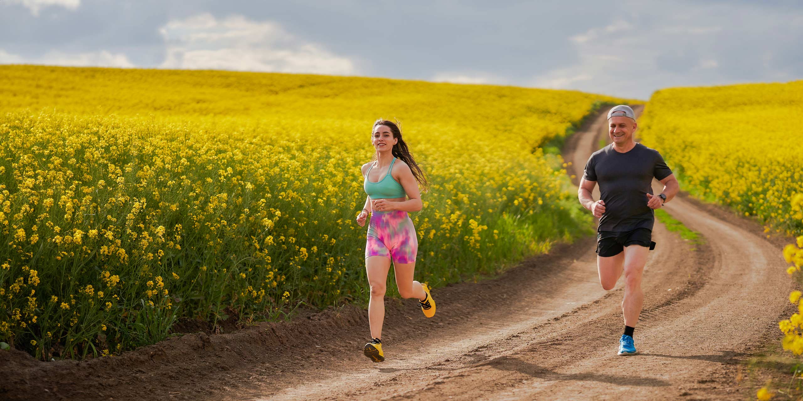 Ein Mann und eine Frau joggen auf einem Feldweg neben einem leuchtend gelben Rapsfeld.